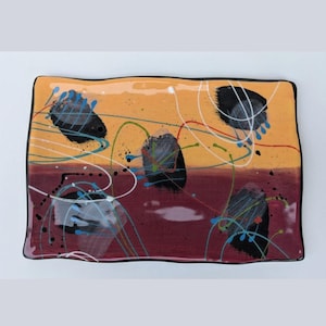 May include: Rectangular ceramic serving dish with an abstract design. The dish features a gradient of colors, including orange, burgundy, and black. Splatters of blue, white, and green paint add to the artistic aesthetic. The edges are outlined in black.