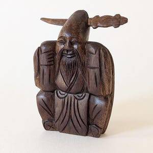 May include: A carved wooden figurine of a seated, smiling elder with a long beard and a pointed hat. The figure is a dark brown color and has a detailed carving of the face, clothing, and a decorative element extending from the hat.
