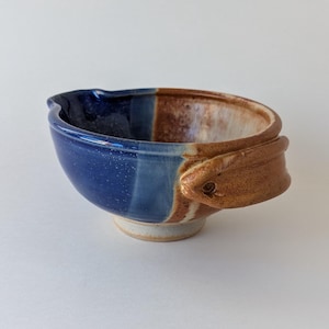 May include: A small, handmade ceramic bowl with a blue and brown glaze. The bowl has a unique, asymmetrical shape and a handle on one side.