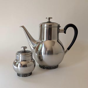 May include: A polished stainless steel teapot and a matching lidded sugar bowl. Both pieces feature a dark, intricate floral pattern band at the base and on the lid knobs. The teapot has a long, curved spout and a black handle for easy pouring.