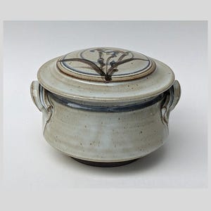 May include: A ceramic lidded casserole dish with a light gray glaze. The lid features a floral design in brown and blue. The dish has two handles and a dark brown base. The overall design is simple and rustic.