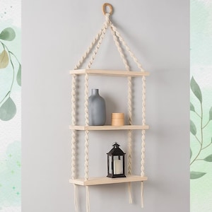 May include: A three-tiered hanging shelf made of light wood and cream-colored rope. The shelf holds a gray vase, a stack of wooden rings, and a black lantern with a white candle. The shelf is suspended from a wooden ring.