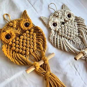 May include: Two macrame owl wall hangings, one in a mustard yellow color and the other in a natural white color. Both owls have brown glass eyes and a wooden dowel attached to the bottom.