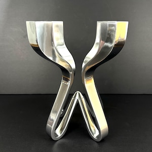 May include: Two polished silver candle holders with a distinctive, curved design. The candle holders have a modern, sculptural appearance, with a base that tapers to a point. The overall shape is symmetrical and elegant, suitable for home decor.