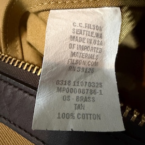 May include: A white fabric product tag with black text reading C.C. FILSON SEATTLE, WA MADE IN USA OF IMPORTED MATERIALS FILSON.COM RN 39126 0318 11070325 MP00008786-1 OS - BRASS TAN 100% COTTON, attached to a tan canvas item with a brass zipper.