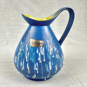 May include: A blue ceramic pitcher with a curved handle and a wide spout. The pitcher has a textured surface with white and light blue drip patterns. The interior of the spout is yellow. A label reads "Carstens Tonkrug Qualitat West-Germany."