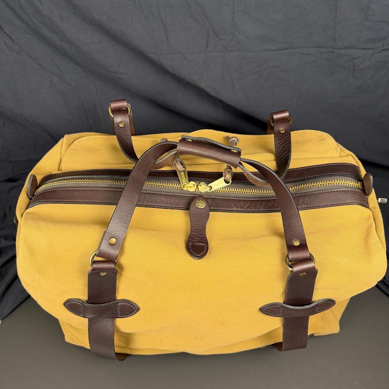 May include: A mustard yellow canvas duffel bag featuring dark brown leather handles, trim, and reinforced strap attachments. The bag includes a top zipper closure with two gold-toned metal pulls, a leather handle wrap, and brass-colored hardware.