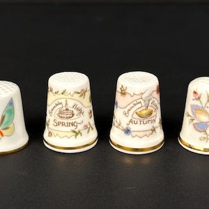 Puede incluir: Cuatro dedal de cerámica blanca con ribete dorado, cada uno con una estación diferente. Los dedales representan una mariposa, un pastel de cumpleaños, un tazón de fruta y una mariposa. El texto en los dedales dice "Brambly Hedge Spring", "Brambly Hedge Autumn" y "Brambly Hedge".