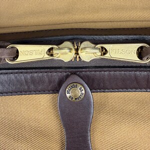 May include: A tan canvas bag featuring two gold-tone metal zipper pulls embossed with the text FILSON. A dark brown leather trim runs horizontally below the zipper, with a leather tab featuring a gold-tone snap button embossed with C.C. FILSON CO. SEATTLE.