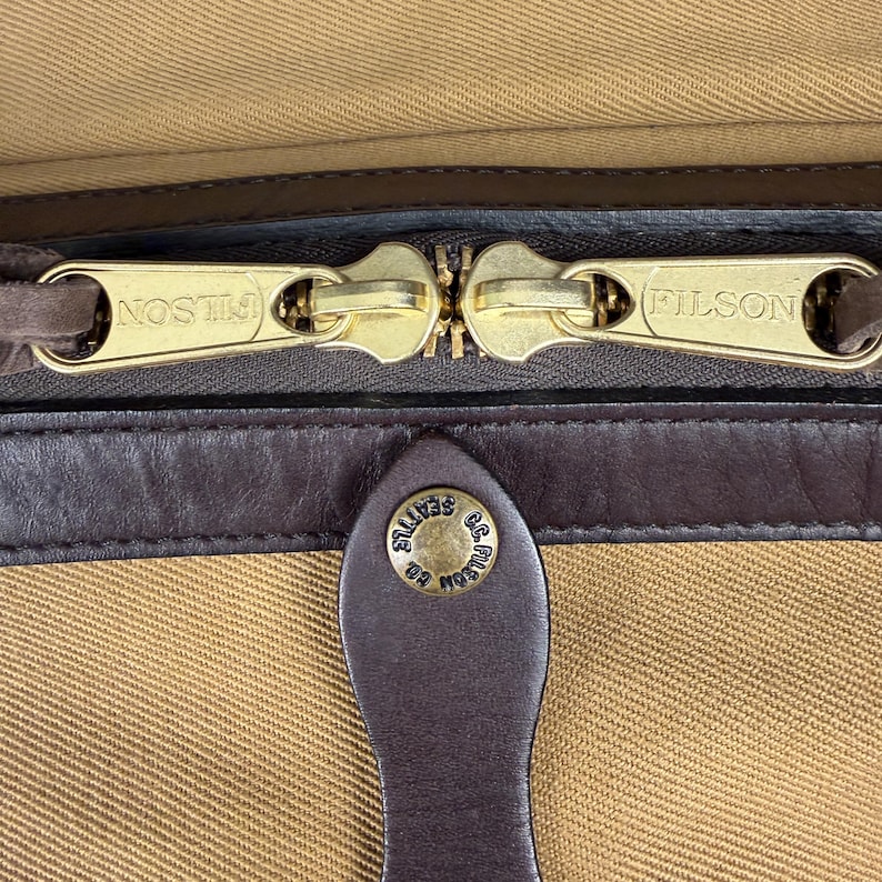 May include: A tan canvas bag featuring two gold-tone metal zipper pulls embossed with the text FILSON. A dark brown leather trim runs horizontally below the zipper, with a leather tab featuring a gold-tone snap button embossed with C.C. FILSON CO. SEATTLE.