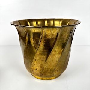 May include: A vintage brass planter with a fluted, spiral design. The planter has a wide, scalloped rim and a slightly flared base. The brass has a warm, aged patina, giving it a classic, antique look. The planter is suitable for indoor or outdoor use.