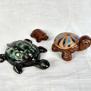 Puede incluir: Tres figuras de tortugas de cerámica. Una es verde oscuro y negra, otra marrón con detalles azules y beige, y la más pequeña es marrón liso. Las tortugas son decorativas y probablemente para exhibición.