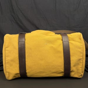 May include: A mustard yellow canvas duffel bag featuring two vertical dark brown leather straps that wrap around the body of the bag. The bag has a rectangular shape with rounded edges and a wide brown fabric shoulder strap attached to the side.