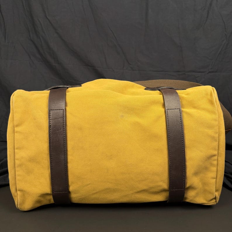 May include: A mustard yellow canvas duffel bag featuring two vertical dark brown leather straps that wrap around the body of the bag. The bag has a rectangular shape with rounded edges and a wide brown fabric shoulder strap attached to the side.