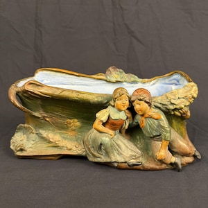 May include: A ceramic planter featuring a sculpted scene of two children sitting together. The exterior is finished in muted green and brown tones with floral relief details, while the interior basin is glazed in a pale, light blue color.