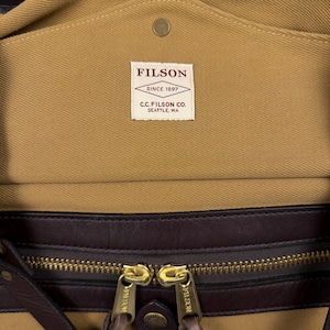 May include: A tan canvas bag featuring a dark brown leather trim and a brass zipper with two pulls. A white rectangular label is stitched to the interior, displaying the text FILSON SINCE 1897 C.C. FILSON CO. SEATTLE, WA in dark red lettering.