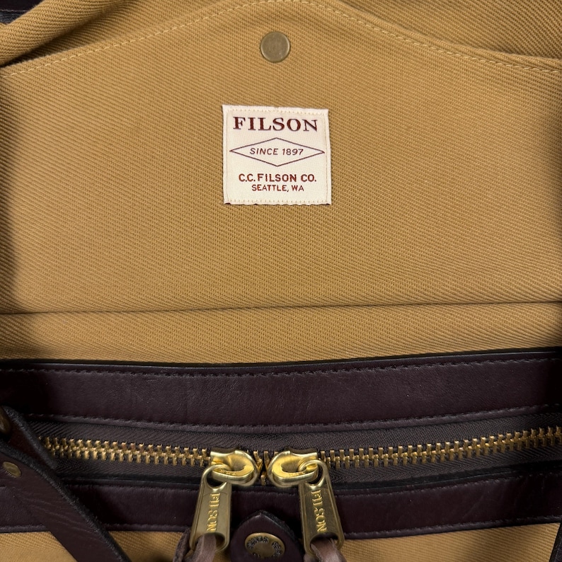 May include: A tan canvas bag featuring a dark brown leather trim and a brass zipper with two pulls. A white rectangular label is stitched to the interior, displaying the text FILSON SINCE 1897 C.C. FILSON CO. SEATTLE, WA in dark red lettering.