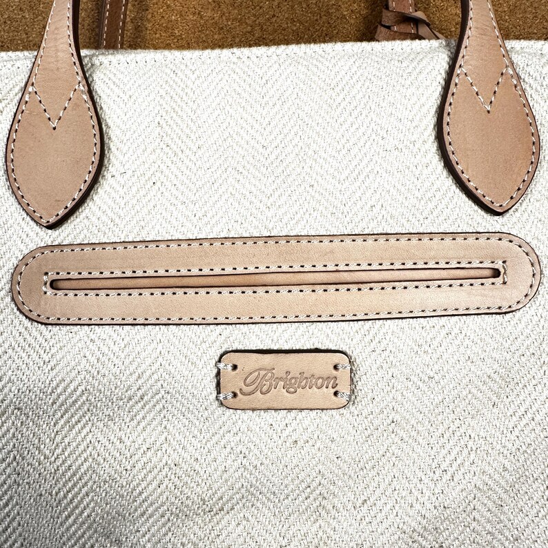 Puede incluir: Primer plano de un bolso tote beige y marr&oacute;n claro. El bolso presenta un patr&oacute;n de espiga, con asas y detalles de cuero marr&oacute;n claro. El nombre de la marca "Brighton" se muestra en una peque&ntilde;a etiqueta de cuero.