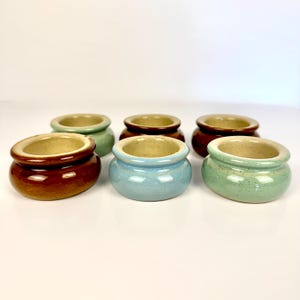 May include: Six small ceramic pots in shades of brown, blue, and green. Each pot has a rounded shape with a contrasting rim and a glossy finish. The pots are displayed on a white surface. The pots are suitable for small plants.