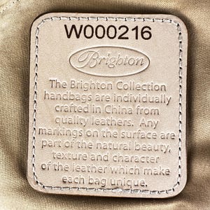 Puede incluir: Una etiqueta rectangular de cuero beige claro con el texto en relieve "Brighton" y el n&uacute;mero "W000216". La etiqueta incluye el texto: "Los bolsos Brighton Collection est&aacute;n hechos individualmente en China con cueros de calidad."