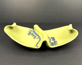 May include: A light green ceramic dish with a unique, curved design. The dish features a central raised section and two flared ends. Blue floral and dot patterns decorate the surface, adding a decorative touch to the piece.