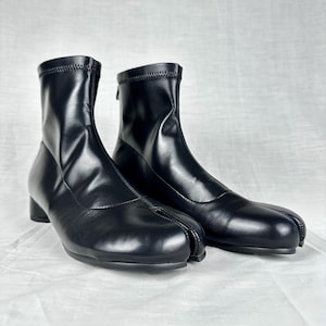 May include: A pair of black split-toe ankle boots. The boots have a shiny finish and a low heel. The design features a split between the big toe and the other toes, and a zip at the back. The boots are approximately 15 cm high.
