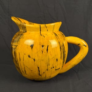 Peut inclure: Un pichet en bois arrondi présentant une finition jaune vif avec des stries de grain noires marquées et des motifs naturels. Le récipient possède un bec verseur large et incurvé, une poignée latérale robuste et une surface polie et lisse.