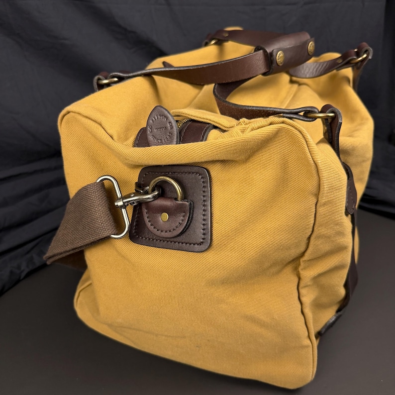 May include: A mustard yellow canvas duffel bag featuring dark brown leather trim, handles, and reinforced corners. The bag includes a detachable brown fabric shoulder strap with metal hardware, a zippered top closure, and a branded leather zipper pull.