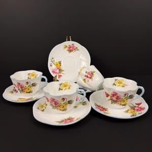 May include: A floral tea set with white porcelain cups and saucers. The cups have light blue handles and rims, and feature a design in yellow, pink, and purple. The set includes a serving plate.