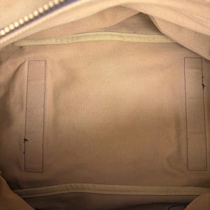 May include: The interior of a bag featuring a light beige fabric lining. The base of the bag is rectangular with two vertical, rectangular fabric panels stitched onto the bottom surface. A zipper track is visible along the top edge of the opening.