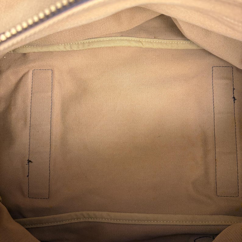 May include: The interior of a bag featuring a light beige fabric lining. The base of the bag is rectangular with two vertical, rectangular fabric panels stitched onto the bottom surface. A zipper track is visible along the top edge of the opening.