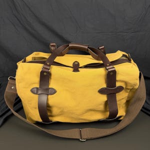 May include: A mustard yellow canvas duffel bag featuring dark brown leather straps, brass-toned hardware, a top zipper closure, and a wide adjustable brown fabric shoulder strap. The bag has two leather handles and reinforced leather accents.