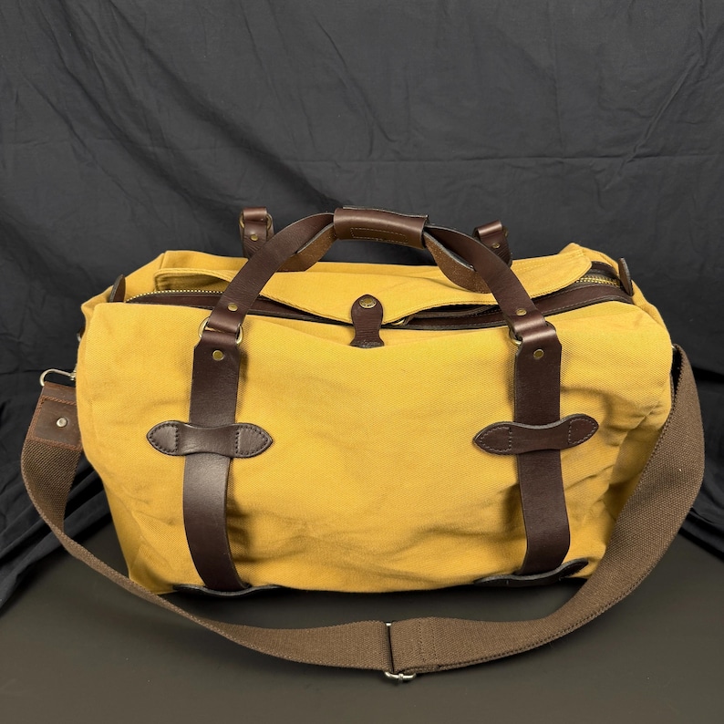 May include: A mustard yellow canvas duffel bag featuring dark brown leather straps, brass-toned hardware, a top zipper closure, and a wide adjustable brown fabric shoulder strap. The bag has two leather handles and reinforced leather accents.