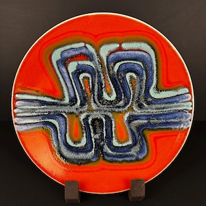 Puede incluir: Un plato redondo de cerámica con un vibrante fondo naranja. El plato presenta un diseño central de formas abstractas azules, turquesas y grises. El plato se exhibe sobre una superficie oscura.