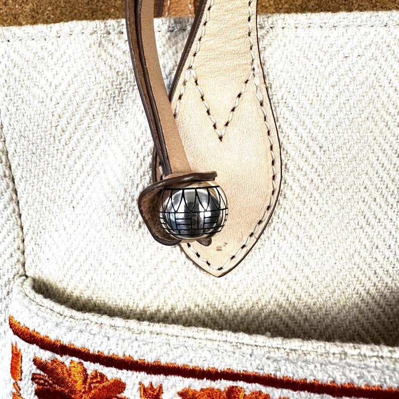 Puede incluir: Primer plano de un bolso tote color crema con un patr&oacute;n de espiga. El bolso presenta un asa de cuero marr&oacute;n y una esfera decorativa de metal plateado. La parte inferior del bolso tiene bordados florales naranjas.