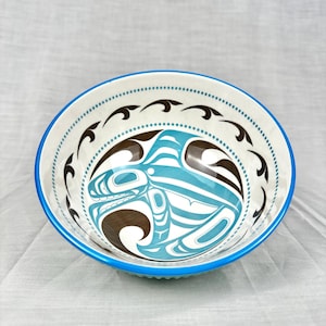 Op de afbeelding: Een keramische kom met een blauwe rand en een witte binnenkant. De kom heeft een centraal ontwerp van een gestileerd blauw en bruin wezen, omgeven door een stippellijn en zwarte gebogen vormen. De kom wordt waarschijnlijk gebruikt om voedsel te serveren.