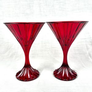 May include: Two ruby red martini glasses. Each glass features a conical bowl with vertical fluting and a stem that flares into a decorative base. The glasses are made of a transparent material.