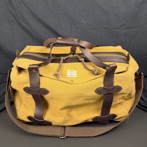 May include: A mustard yellow canvas duffel bag featuring dark brown leather straps, brass hardware, and a top zipper closure. A white rectangular label with the text FILSON is centered on the front. The bag includes a wide brown shoulder strap.
