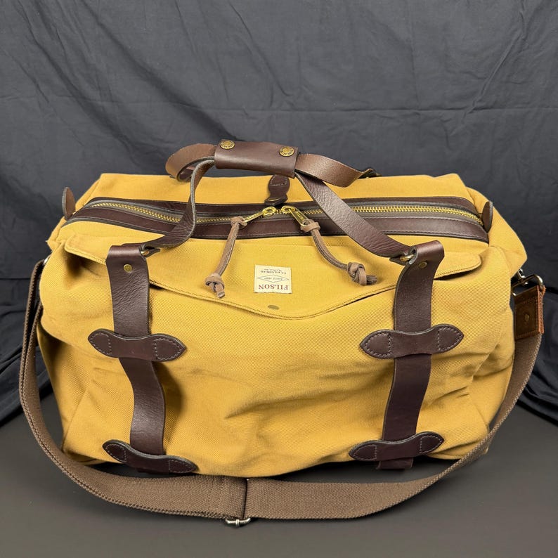 May include: A mustard yellow canvas duffel bag featuring dark brown leather straps, brass hardware, and a top zipper closure. A white rectangular label with the text FILSON is centered on the front. The bag includes a wide brown shoulder strap.