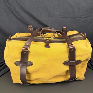 May include: A mustard yellow canvas duffel bag featuring dark brown leather straps and trim. The bag includes a top zipper closure with two brass-toned metal pulls, leather handle wraps, and reinforced leather accents secured with brass rivets.