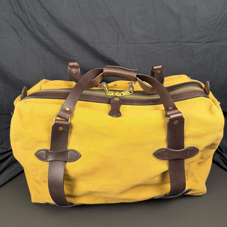 May include: A mustard yellow canvas duffel bag featuring dark brown leather straps and trim. The bag includes a top zipper closure with two brass-toned metal pulls, leather handle wraps, and reinforced leather accents secured with brass rivets.