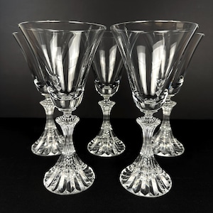 May include: A collection of six clear crystal wine glasses. Each glass has a flared bowl, an ornate stem, and a detailed base. The glasses are displayed on a dark surface, highlighting their elegant design.