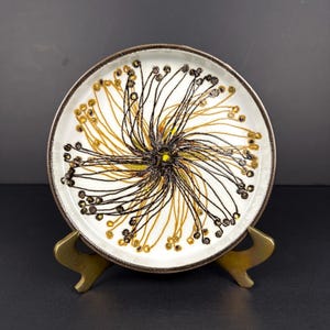 Può includere: Un piatto in ceramica bianca con bordo marrone e un disegno centrale di linee nere e dorate che si irradiano da un centro giallo. Il piatto è esposto su un supporto in metallo color oro, su sfondo scuro.