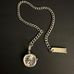 May include: Silver pocket watch with a chain, displaying a cartoon character on the watch face. The watch has a silver bezel and a chain with a rectangular tag that reads "Trecceto". The watch is on a black surface.