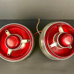 Puede incluir: Par de luces traseras de coche vintage. Cada luz presenta una lente roja brillante con un bisel cromado. El diseño incluye una lente roja central y tres soportes de montaje plateados. Se ven cables.