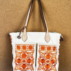 Puede incluir: Bolso tote blanco con bordado floral naranja en los bolsillos delanteros. El bolso tiene asas y detalles de cuero marr&oacute;n claro. La cremallera es plateada. El bolso est&aacute; hecho de una tela texturizada.