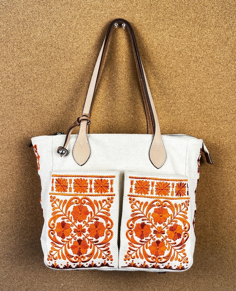 Puede incluir: Bolso tote blanco con bordado floral naranja en los bolsillos delanteros. El bolso tiene asas y detalles de cuero marr&oacute;n claro. La cremallera es plateada. El bolso est&aacute; hecho de una tela texturizada.