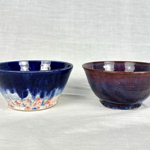 Op de afbeelding: Twee handgemaakte keramische kommen. Eén kom is diepblauw met witte en rode accenten. De andere kom heeft een verloop van paars en bruin. Beide kommen hebben een glanzende afwerking en staan tegen een witte achtergrond.