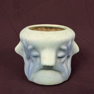 May include: A light blue ceramic planter with a crackled glaze finish. The exterior features a sculpted, symmetrical face design with closed eyes and a prominent nose. The vessel has a wide, circular opening at the top and a smooth base.