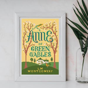 May include: Framed cross-stitch art featuring the book cover of "Anne of Green Gables." The design includes the title in teal and white, with a yellow background. The artwork depicts trees, a house, and the author's name.
